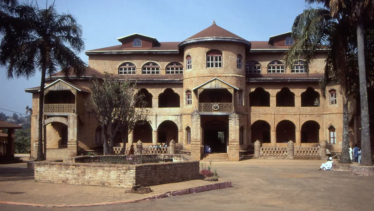 The arcaded brick facade of the Sultan's palace at Foumban, framed by palm trees under a blue sky.