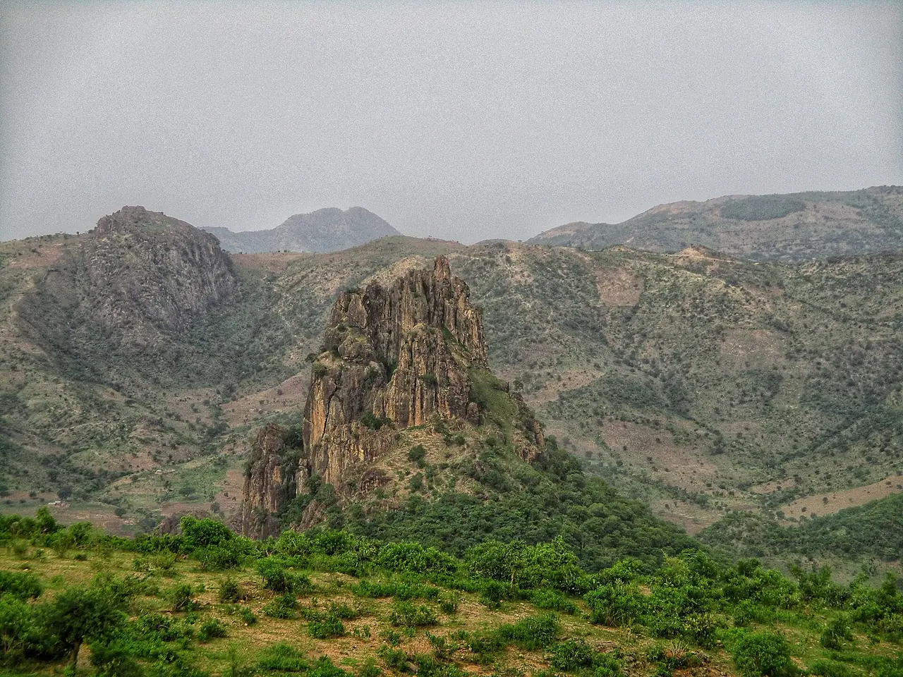 A jagged volcanic rock spire rising above green, scrub-covered hills in the Kapsiki mountains.
