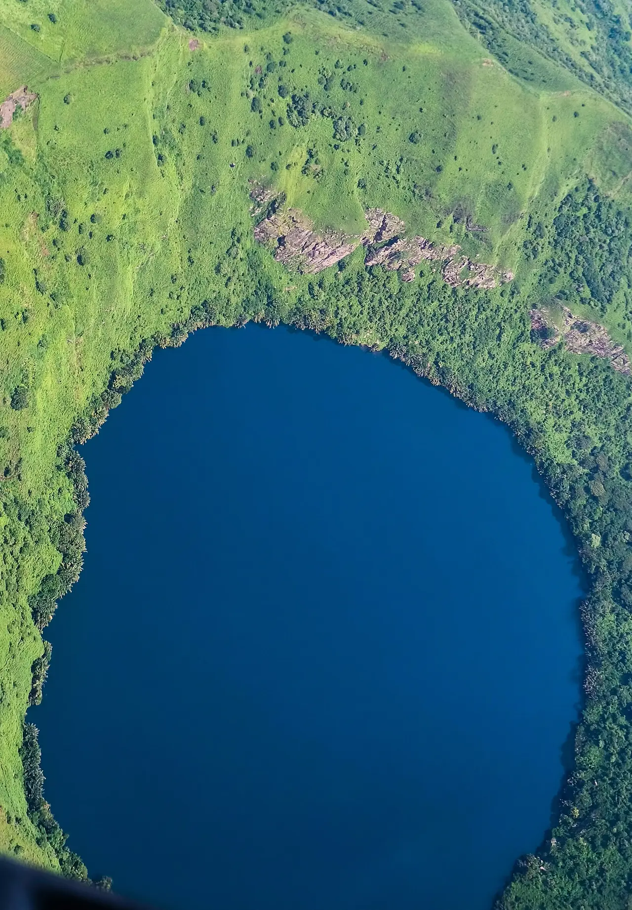 Aerial view of Lake Kuk, a deep blue crater lake ringed by forest and green grassland.